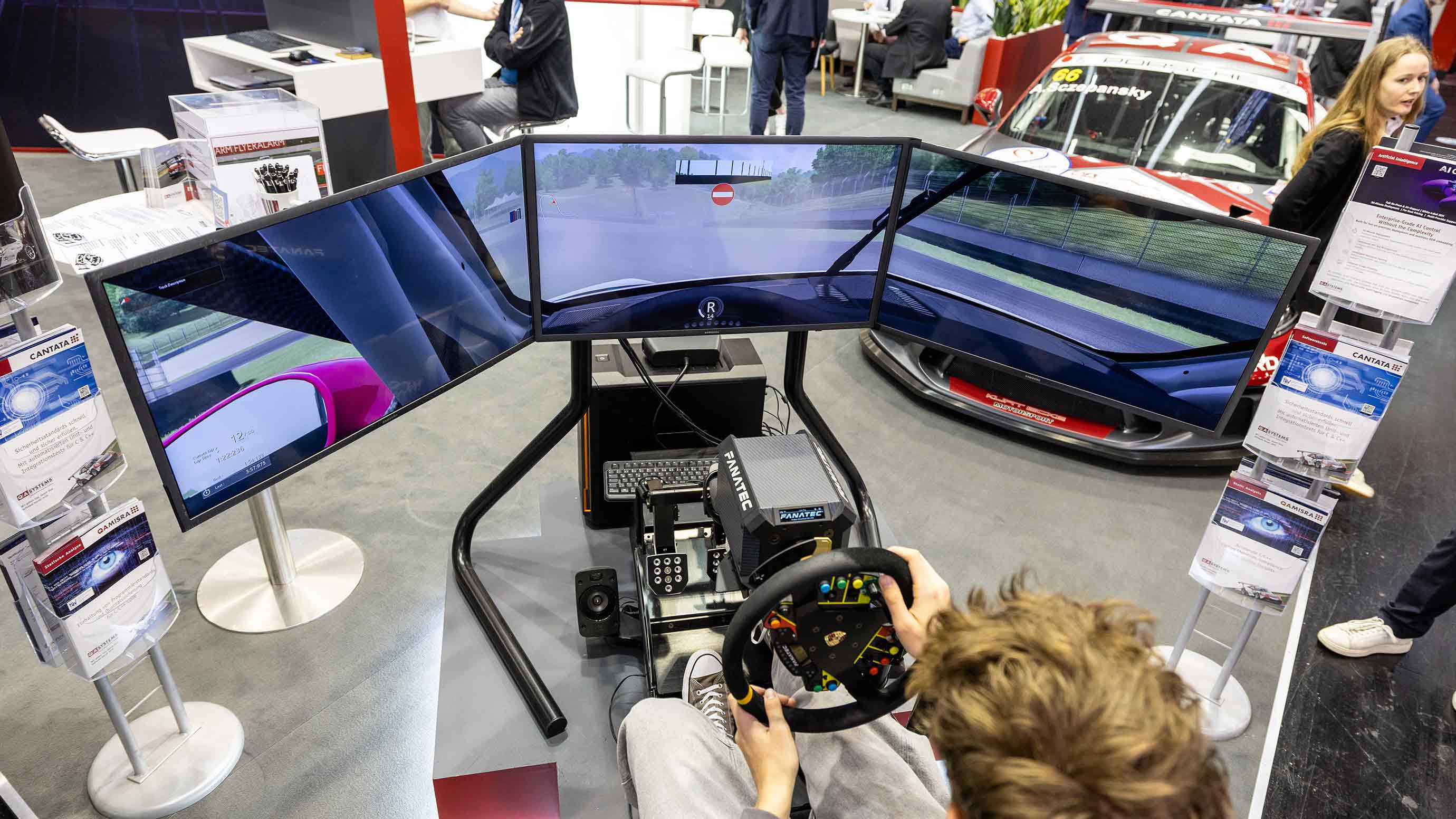 Person testing a racing simulator with steering wheel, pedals, and multiple screens at a trade fair.