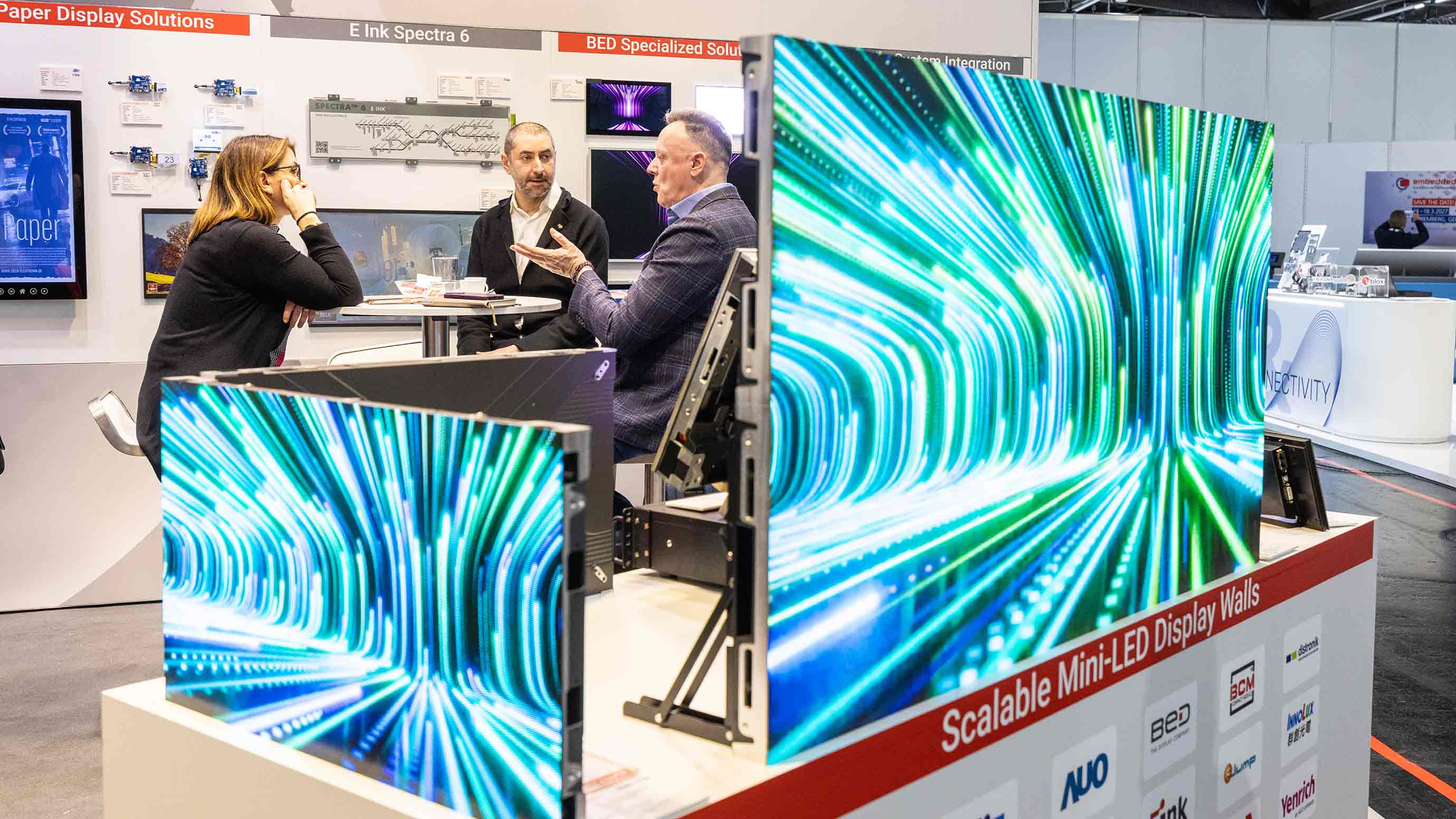 Discussion at an exhibition booth featuring large mini LED displays showing abstract light graphics.